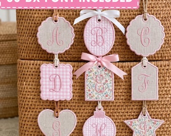 May include: A collection of nine fabric ornaments featuring embroidered letters A through I. The ornaments vary in shape, including scalloped circles, squares, a heart, and a star, with pink, beige, and floral patterns, some accented with pink satin bows.