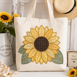 Peut inclure: Un sac fourre-tout en toile couleur crème orné d'un grand motif de tournesol central. Le tournesol possède un cœur brun foncé et des pétales jaunes aux motifs variés, incluant du vichy et des fleurs, avec deux feuilles vert sauge à la base.