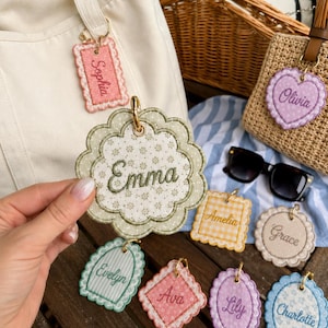 May include: A collection of embroidered fabric bag tags in various shapes and colors, each featuring a unique name stitched in cursive script. The tags include names such as Emma, Sophia, Olivia, Amelia, Grace, Evelyn, Ava, Lily, and Charlotte.
