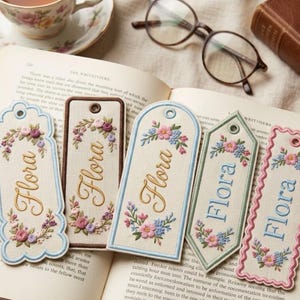 May include: Five rectangular fabric bookmarks with embroidered floral wreaths and the name Flora in cursive script. Each bookmark features a unique edge shape, including scalloped, rounded, and pointed designs, with a small eyelet at the top.