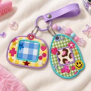May include: A keychain featuring two embroidered patches shaped like handheld digital pets. One is a yellow rectangle with a blue gingham screen, the other an oval with a green gingham pattern and leopard print screen, accented with beads.