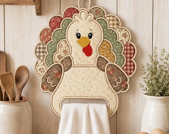 May include: A fabric turkey-shaped kitchen towel holder featuring a cream-coloured body with a floral pattern, a yellow beak, and a red wattle. The turkey has a scalloped tail made of various patterned fabric patches in red, green, and brown tones.