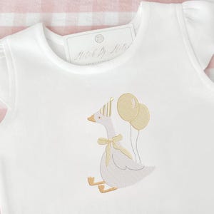 May include: White onesie with a whimsical embroidered goose wearing a party hat and bow tie, holding two balloons. The goose is in shades of gold and tan. The onesie has short sleeves and a tag that reads "Match By Style".