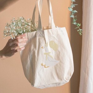 May include: Cream-colored tote bag with a whimsical embroidered design of a goose wearing a party hat and holding yellow balloons. The goose has a yellow bow. The bag is filled with white flowers.