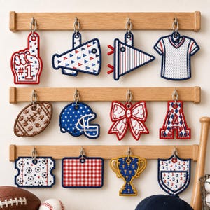 May include: A wooden rack with three horizontal bars holding twelve hanging ornaments featuring sports-themed designs including a foam finger, megaphone, pennant, jersey, football, helmet, bow, letter A, soccer ball, trophy, and shield.