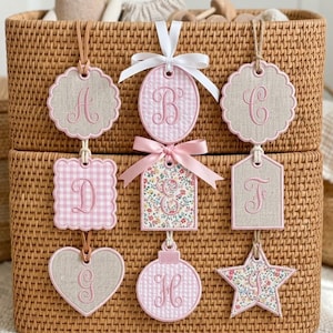 Puede incluir: Una colección de etiquetas decorativas en varias formas, incluyendo un corazón, una estrella y un óvalo, con letras bordadas. Las etiquetas presentan patrones rosas y florales, con bordes festoneados y cintas. Cuelgan de una cesta tejida.