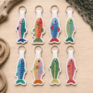 Puede incluir: Ocho llaveros de peces bordados dispuestos en dos filas. Cada llavero cuenta con una lengüeta de cuero blanco con un anillo de metal plateado y un diseño de pez colorido con escamas, aletas y un ojo cosidos.