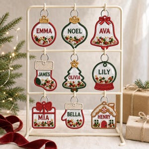 Op de afbeelding: Negen geborduurde kerstornamenten hangend aan een crèmekleurige metalen standaard. Elk ornament heeft een doorzichtig midden gevuld met kleurrijke kralen en pailletten, gepersonaliseerd met de namen Emma, Noel, Ava, James, Olivia, Lily, Mia, Bella en Henry.