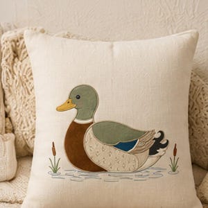 Puede incluir: Un cojín cuadrado de color blanco roto con un diseño bordado de un pato azulón. El pato tiene la cabeza verde, el pecho marrón y alas estampadas en tonos beige, azul y negro. Pequeñas espadañas y ondas de agua bordadas rodean al pato.