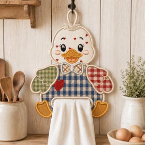 May include: A hanging kitchen towel holder shaped like a duck with a cream-colored head, a yellow beak, and a blue gingham body. The duck features a small bowtie, a red heart patch, and wings with green and red checkered patterns.