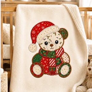 May include: A cream-colored fleece blanket featuring a central embroidered teddy bear wearing a red Santa hat with white snowflakes, a green and red plaid scarf, and holly leaf accents. The bear has a light tan face and dark brown eyes.