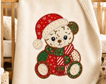 Op de afbeelding: Een crèmekleurige fleece deken met een centrale geborduurde teddybeer die een rode kerstmuts met witte sneeuwvlokken, een rood-groene sjaal met ruitpatroon en hulstblaadjes draagt. De beer heeft een lichtbruin gezicht en donkerbruine ogen.