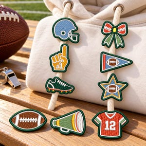 May include: A cream-colored hoodie with two drawstrings decorated with embroidered patches featuring football-themed icons including a helmet, a foam finger, a cleat, a bow, a pennant, a star, a football, a megaphone, and a jersey with the number 12.