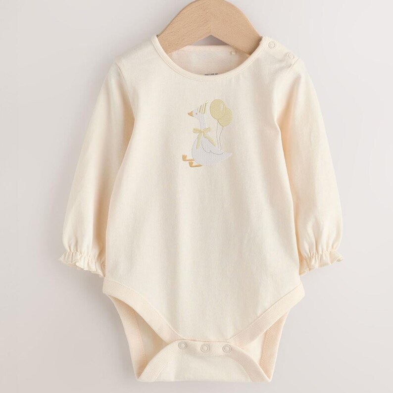 May include: Cream-colored baby bodysuit with long sleeves and a round neckline. The bodysuit features a whimsical embroidered design of a goose holding balloons. The sleeves have a ruffled cuff. The bodysuit has snap closures at the bottom and on the shoulder.