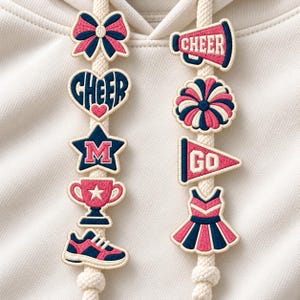 May include: A cream-colored hoodie featuring two thick drawstrings adorned with embroidered patches. The left string displays a bow, a heart with the word CHEER, a star with the letter M, a trophy, and a sneaker. The right string features a megaphone with the word CHEER, a pom-pom, a pennant with the word GO, and a pleated skirt.