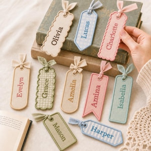 May include: A collection of rectangular fabric bookmarks featuring embroidered names including Olivia, Lucas, Charlotte, Evelyn, Grace, Amelia, Amina, Isabella, Mason, and Harper. Each bookmark has a fabric ribbon tie at the top and various edge shapes.