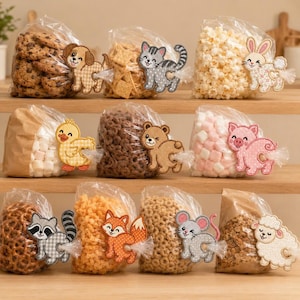 Op de afbeelding: Twaalf doorzichtige plastic zakjes gevuld met diverse snacks, waaronder koekjes, crackers, popcorn, marshmallows en ontbijtgranen. Elk zakje is verzegeld met een decoratieve geborduurde dierenpatch, met een hond, kat, konijn, eend, beer, varken, wasbeer, vos, muis en schaap.