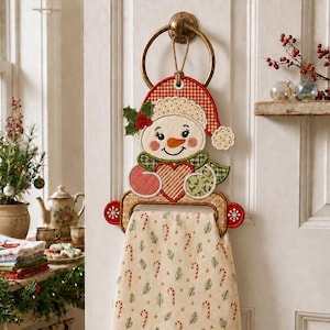 May include: A decorative fabric snowman towel holder hanging from a brass ring. The snowman wears a red and white checkered hat, a green scarf, and holds a red and white striped heart. A cream-colored towel with a candy cane and pine print hangs below.