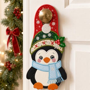 May include: A fabric door hanger shaped like a penguin wearing a red and green hat with white snowflake patterns, a light blue scarf with white dots, and orange feet. The penguin has a white belly, black body, and orange beak.
