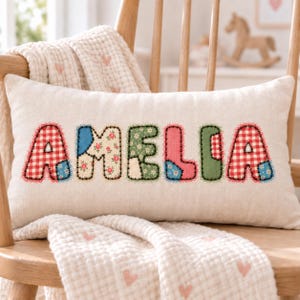 Puede incluir: Un cojín rectangular de color blanco roto que presenta el nombre AMELIA aplicado en letras grandes cosidas. Cada letra está confeccionada con diferentes telas estampadas, incluyendo cuadros vichy rojos, motivos florales y parches lisos verdes y rosas.