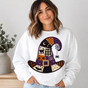 May include: A white long-sleeve sweatshirt featuring a large graphic of a witch hat. The hat is designed with a patchwork pattern including orange, black, and purple sections, decorated with motifs of a black cat, pumpkins, a bat, and a spider.