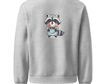 May include: A light gray sweatshirt featuring a cartoon raccoon holding a blue bowl filled with colorful candies. The raccoon has a black mask, gray fur, and a striped tail. The sweatshirt has a crew neck and long sleeves.
