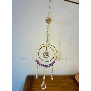 Puede incluir: Un atrapasoles dorado con un colgante de árbol de la vida, virutas de amatista y detalles de cristal transparente. El atrapasoles está suspendido por una cadena dorada y presenta el texto "@kalai handmade".
