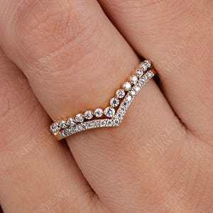 May include: A V-shaped chevron ring crafted from yellow gold, featuring two distinct bands. The upper band is set with a series of round, bezel-set white stones, while the lower band is pavé-set with smaller, shimmering white stones.