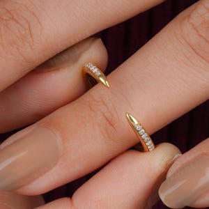 May include: A gold open-style ring featuring two pointed ends, each accented with a row of small, clear pavé-set stones. The ring is designed with a sleek, polished finish and a tapered silhouette that narrows toward the center of the band.