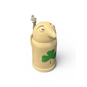 May include: A cream-colored ceramic beer stein featuring a lid shaped like a face with a prominent nose, two eyes, and orange eyebrows. A metallic thumb lift is attached to the lid. A green three-leaf clover is printed on the front body.