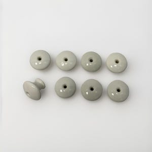 May include: Eight light gray ceramic cabinet knobs. Seven are round with a central hole, and one is a knob with a rounded base and a cylindrical handle. The knobs are arranged on a white background.