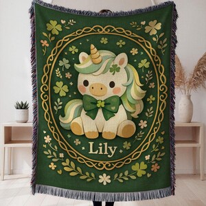 May include: Green woven throw blanket with a cartoon unicorn design. The unicorn has a golden horn, green bow, and rainbow mane, surrounded by shamrocks and the name "Lily". The blanket has a fringed edge.