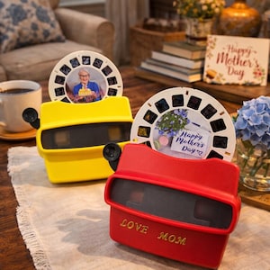 Puede incluir: Dos visores de estilo retro, uno amarillo y otro rojo, sobre una superficie de madera. El visor rojo tiene impreso "LOVE MOM". Cada visor contiene una diapositiva circular con imágenes. Una diapositiva dice "Happy Mother's Day".