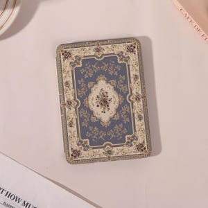 May include: A rectangular box with a vintage rug design. The box has a central medallion with a bouquet of flowers in burgundy and cream, set against a dusty blue background. The border is cream with floral accents. The box is approximately 15 cm x 10 cm.