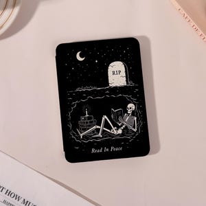 May include: A black rectangular e-reader case featuring a white line art illustration of a skeleton reading a book inside an underground cavern beneath a tombstone marked RIP, with the text Read In Peace printed at the bottom of the cover.