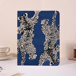 Può includere: Un taccuino blu con un motivo animalier in bianco e nero. La copertina presenta un motivo ripetuto di figure di animali stilizzate. Il taccuino è rettangolare e sembra chiuso.
