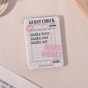 May include: A white rectangular notepad with the words "GUEST CHECK" at the top. The notepad has lines and the phrases "make love, make out, make art" and "MAKE MONEY" in pink. The check number is 037594.