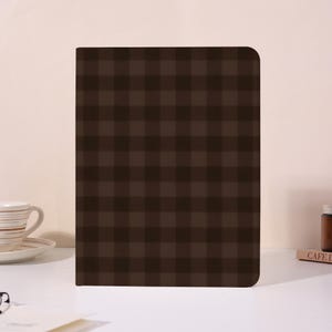 May include: A rectangular notebook with a dark brown and black plaid pattern. The cover features a consistent grid design with alternating dark squares. The notebook has rounded top corners and a flat base, standing upright on a smooth surface.