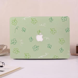 May include: A light green laptop case featuring a pattern of green shamrocks and white swirls. The Apple logo is visible in the center. The case is on a white surface.