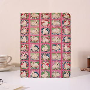 May include: A rectangular notebook featuring a repeating grid pattern of white rabbits. Each rabbit is enclosed in a square or circular frame with colorful decorative borders in shades of pink, green, blue, and yellow against a bright pink background.