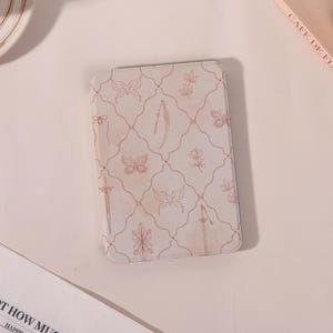 May include: A rectangular, off-white case with a repeating pattern of butterflies, flowers, and swords in a soft rose color. The case has rounded corners and a smooth surface. The design is symmetrical and delicate.