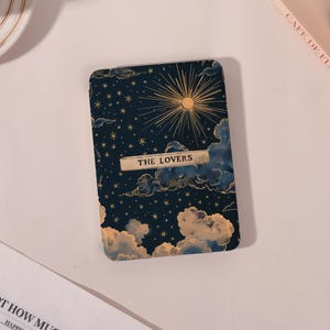 May include: A tarot card with a night sky design, featuring gold stars, a sunburst, and the text "THE LOVERS". The card has a dark blue background with fluffy clouds at the bottom. The card is rectangular with rounded corners.