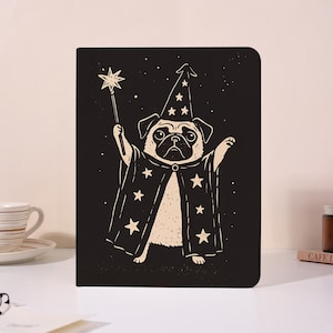 Puede incluir: Un cuaderno negro con una ilustración de un pug vestido de mago. El pug lleva un sombrero puntiagudo y una capa con estrellas, sosteniendo una varita en forma de estrella. El fondo es negro con estrellas blancas.