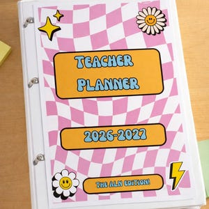 K&ouml;nnte beinhalten: Wei&szlig;er Ordner mit einem rosa-wei&szlig; karierten Design. Auf dem Cover steht "TEACHER PLANNER 2026-2027" in orangefarbenem Text. Dekorative Aufkleber sind ein gelber Stern, ein G&auml;nsebl&uuml;mchen mit l&auml;chelndem Gesicht und ein Blitz. Der Text "THE ALN EDITION!" ist ebenfalls sichtbar.