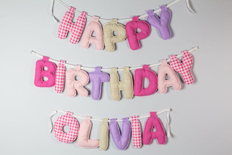 May include: A handmade banner with the words "HAPPY BIRTHDAY OLIVIA" in colorful, fabric letters. The letters are in shades of pink, purple, and beige, with some featuring a gingham pattern. The banner is strung on white string.