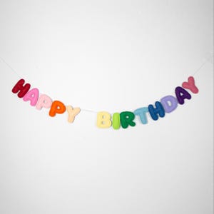 Può includere: Un colorato banner in feltro con la scritta "HAPPY BIRTHDAY". Le lettere sono di vari colori, tra cui rosso, rosa, arancione, giallo, verde, blu e viola, infilate su uno spago bianco su sfondo bianco.