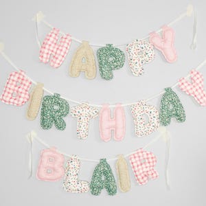 May include: A three-tiered banner with the words "HAPPY BIRTHDAY BLAIR" spelled out in fabric letters. The letters are in various patterns and colors, including pink, green, and beige, strung on white ribbon.