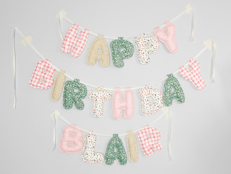 May include: A three-tiered banner with the words "HAPPY BIRTHDAY BLAIR" spelled out in fabric letters. The letters are in various patterns and colors, including pink, green, and beige, strung on white ribbon.