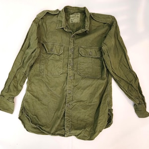May include: An olive green long-sleeved shirt with two chest pockets and button fastenings. The shirt features a traditional collar and shoulder epaulettes. The fabric looks worn and durable. A small label is visible near the collar.