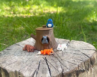 Könnte beinhalten: Eine skurrile Waldszene mit Miniatur-Tonfiguren. Ein Baumstumpf trägt einen braunen baumförmigen Behälter mit einem blauen Vogel, einem grauen Eichhörnchen, einem braunen Bären, einem orangefarbenen Fuchs und einem weißen Kaninchen. Die Szene spielt im Freien.